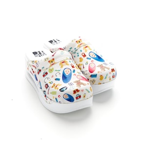 Op de afbeelding: Een paar witte klompen met een kleurrijk patroon van babygerelateerde iconen, waaronder teddyberen, fopspenen en babyflesjes. De klompen hebben een witte platformzool en een zwart logo met de tekst "Dr. Moon".