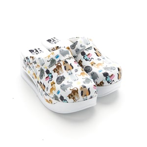 May include: A pair of white clogs with a cat print design. The clogs have a thick white sole and a strap across the top.