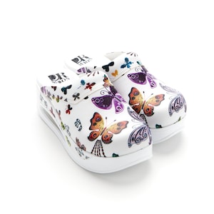 Peut inclure: Sabots blancs avec un imprimé papillon. Les sabots ont une épaisse semelle blanche et un logo "DR. MOON" noir sur le côté.