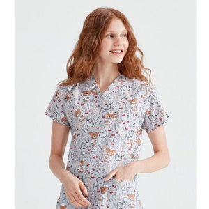May include: A gray medical scrub top with a V-neck and short sleeves. The top features a repeating pattern of teddy bears wearing doctor's hats and holding medical tools.