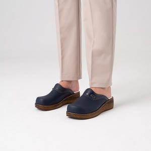 Puede incluir: Zuecos azul marino con suela de madera. Los zuecos tienen una parte superior de cuero texturizado y un pequeño detalle decorativo en el lateral. Los zapatos se usan con pantalones beige.
