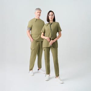 Puede incluir: Dos personas vestidas con uniformes médicos verde oliva. Los uniformes incluyen tops de manga corta y pantalones a juego. Una persona tiene un estetoscopio. Ambos llevan zapatillas blancas. El fondo es blanco liso.
