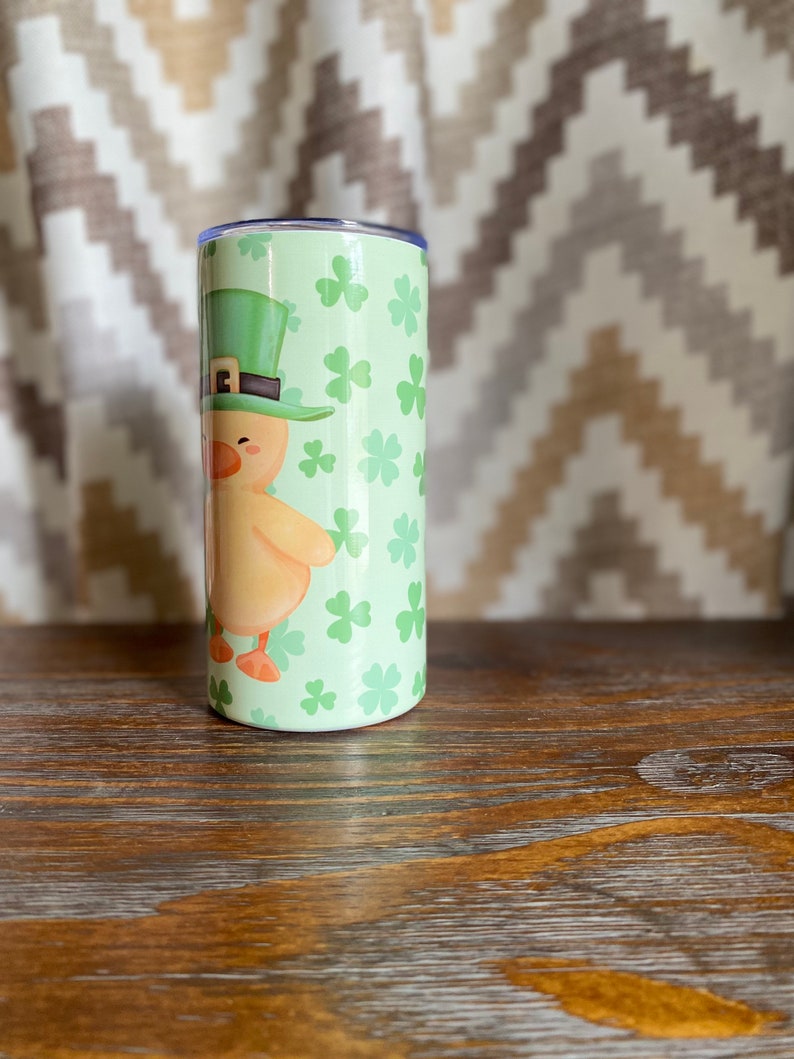 May include: A green and yellow tumbler with a cartoon image of a chick wearing a green leprechaun hat. The tumbler is covered in a pattern of green shamrocks.