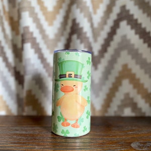 May include: A green and yellow tumbler with a cartoon duck wearing a green leprechaun hat. The tumbler is decorated with shamrocks.