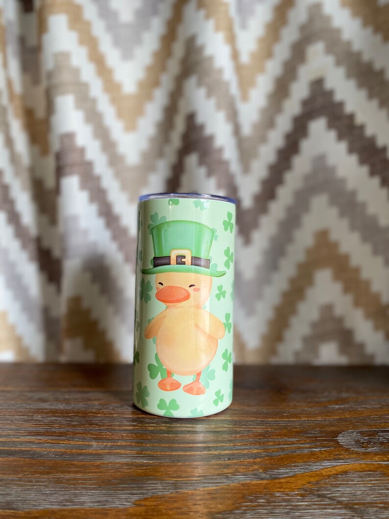 May include: A green and yellow tumbler with a cartoon duck wearing a green leprechaun hat. The tumbler is decorated with shamrocks.