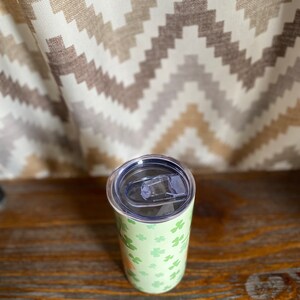 May include: A stainless steel tumbler with a green and white shamrock pattern. The tumbler has a lid with a clear plastic slider.
