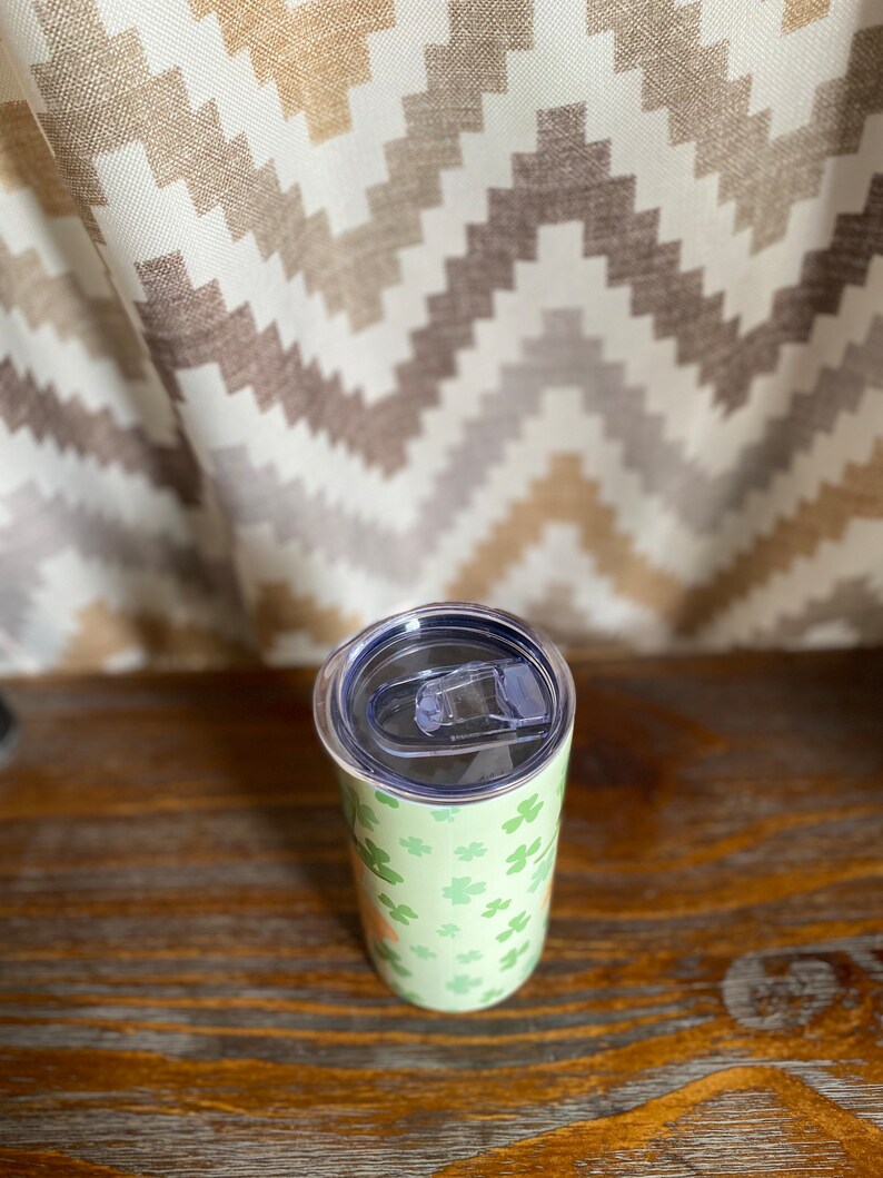 May include: A stainless steel tumbler with a green and white shamrock pattern. The tumbler has a lid with a clear plastic slider.