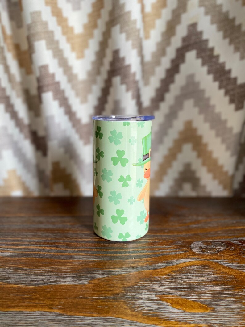May include: A green and white stainless steel tumbler with a shamrock and leprechaun design. The tumbler has a lid and is perfect for holding beverages.