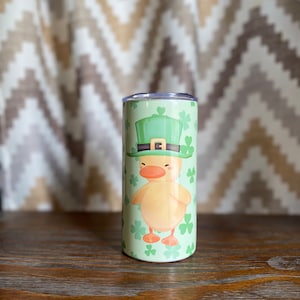 May include: A green and yellow insulated tumbler with a cartoon duck wearing a green leprechaun hat and holding a shamrock. The tumbler is surrounded by green shamrocks.