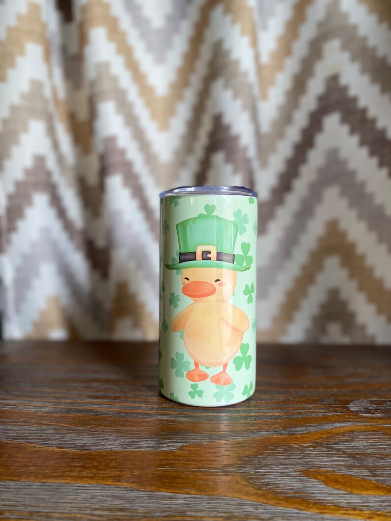 May include: A green and yellow insulated tumbler with a cartoon duck wearing a green leprechaun hat and holding a shamrock. The tumbler is surrounded by green shamrocks.