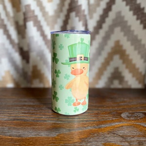 May include: A green and white stainless steel tumbler with a cartoon image of a duck wearing a green leprechaun hat and holding a shamrock. The background is a green shamrock pattern.
