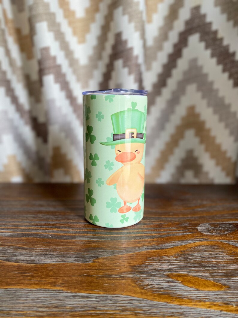 May include: A green and white stainless steel tumbler with a cartoon image of a duck wearing a green leprechaun hat and holding a shamrock. The background is a green shamrock pattern.
