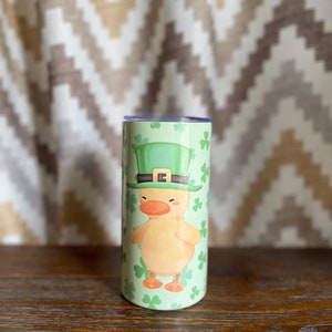 May include: A green and yellow tumbler with a cartoon duck wearing a green leprechaun hat and holding a shamrock. The tumbler is surrounded by shamrocks.