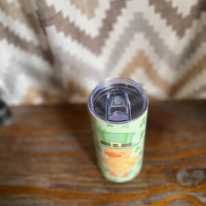May include: A stainless steel tumbler with a green, white, and yellow design of a leprechaun wearing a green hat and holding a pot of gold. The tumbler has a clear lid.