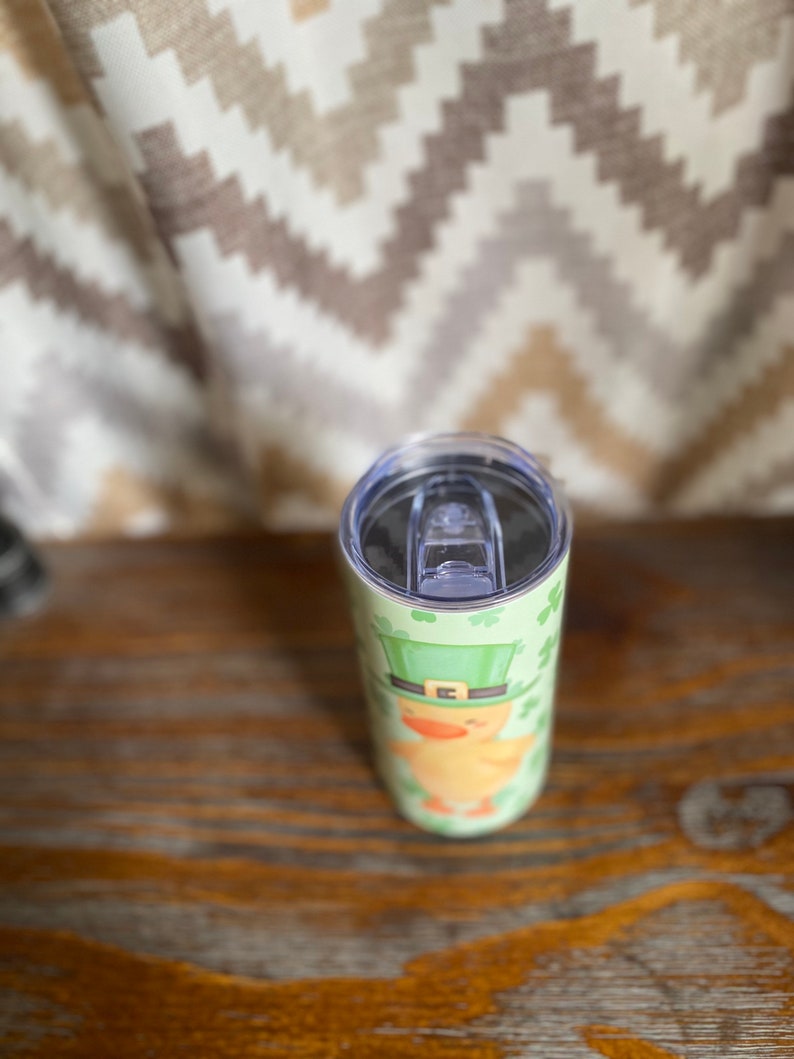 May include: A stainless steel tumbler with a green, white, and yellow design of a leprechaun wearing a green hat and holding a pot of gold. The tumbler has a clear lid.