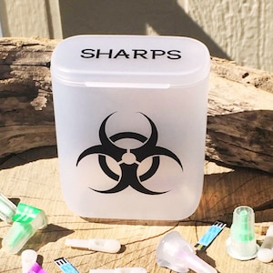 Könnte beinhalten: Ein weißer Plastikbehälter für scharfe Gegenstände mit einem schwarzen Biohazard-Symbol und dem Wort "SHARPS" auf dem Deckel. Der Behälter steht auf einer Holzoberfläche, auf der mehrere medizinische Nadeln und Lanzetten verstreut liegen.