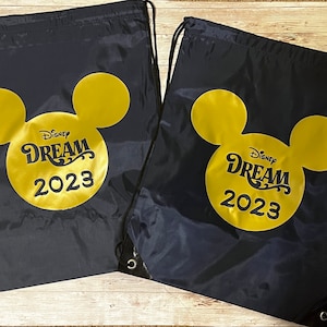 Könnte beinhalten: Zwei schwarze Kordelzugbeutel mit einem goldenen Mickey-Maus-Kopfdesign. Die Beutel tragen den Text "Disney Dream 2023" innerhalb des Mickey-Maus-Kopfes.