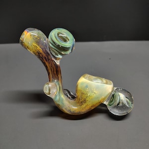 May include: A colorful glass pipe with a curved design. The pipe features a bowl, a stem, and a mouthpiece. The glass has a mix of colors, including yellow, green, brown, and clear. The pipe is designed for smoking.