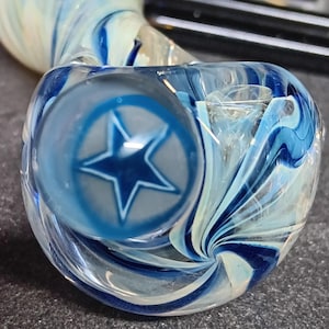 May include: A clear glass pipe with a blue star design. The pipe features a swirling pattern of blue and white, with a bowl-shaped head and a stem. The star is inside a circle on the side of the bowl.