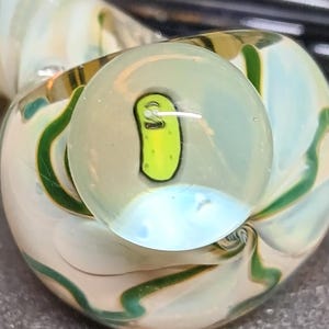 May include: A glass pipe with a light green and white swirl design. The bowl features a cartoon pickle character. The pipe has a rounded shape with green accents and a clear mouthpiece. The design is detailed and artistic.