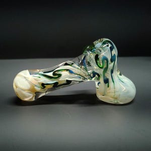 May include: A clear glass pipe with a hammer-like shape. The pipe features swirling patterns of white, green, blue, and yellow. The bowl and mouthpiece are rounded, and the body is elongated.