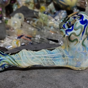 May include: A hand-blown glass pipe with a swirling design of blue, green, and white colors. The pipe has a rounded bowl and a long, slender stem. The glass is translucent, allowing light to pass through.