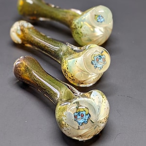 May include: Three glass pipes with a green and yellow swirl design. Each pipe has a round bowl with a cartoon image of a blue Pokemon character.