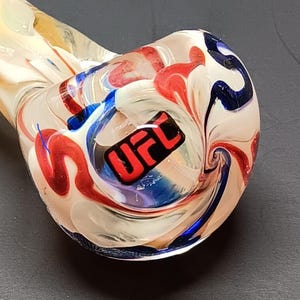 May include: A clear glass pipe with a swirling design in red, white, and blue. The pipe has a rounded bowl with the letters "UFC" in black and red. The stem is a light amber color.