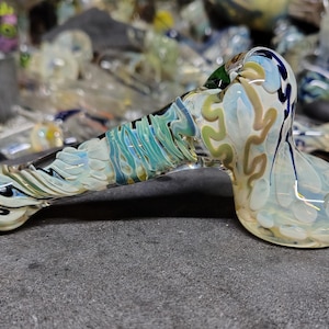 May include: A colorful glass pipe with a rounded bowl and a long, curved stem. The pipe features a mix of colors, including blue, green, white, and brown, with a swirling pattern. The pipe is resting on a dark gray surface.