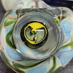 May include: A clear glass smoking pipe with a bowl featuring a yellow and black logo. The pipe has swirls of blue, green, and yellow glass. The logo is a stylized letter "W" inside a circle.