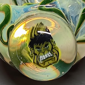 May include: A glass pipe with a Hulk graphic. The pipe features a green and yellow color scheme with a clear bowl. The Hulk's face is visible in the bowl, with a black hairline and angry expression.