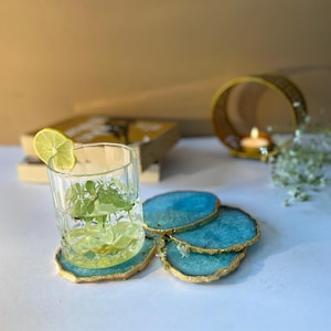 May include: Four blue agate coasters with gold trim, stacked on a white surface. A glass of water with a lime wedge and mint leaves sits on top of the coasters.