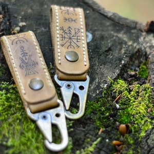 May include: Two tan leather keychains with metal clasps and engraved symbols. The keychains are attached to a wooden surface covered in green moss. The leather has a worn appearance, and the symbols are dark.