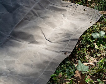 May include: A large, gray canvas tarp with brass grommets and leather straps. The tarp is lying on a bed of green grass and brown leaves.