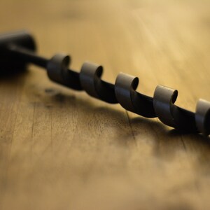 May include: A black metal drill bit with a spiral design. The drill bit is lying on a wooden surface.