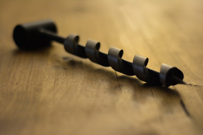 May include: A black metal drill bit with a spiral design. The drill bit is lying on a wooden surface.