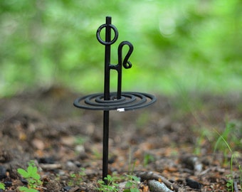May include: A black metal mosquito coil holder with a spiral coil and a decorative top. The holder is standing on a bed of brown leaves and green grass.