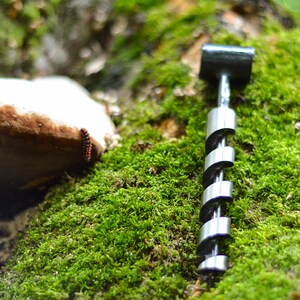 May include: A silver drill bit with a black handle, lying on a bed of green moss. The drill bit is made of metal and has a spiral design. The moss is soft and green, and the background is blurred.