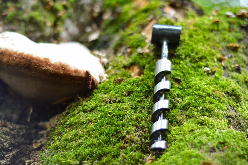 May include: A silver drill bit with a black handle, lying on a bed of green moss. The drill bit is made of metal and has a spiral design. The moss is soft and green, and the background is blurred.