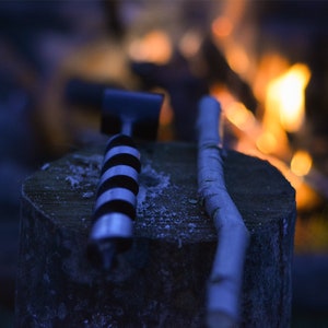 May include: A close-up of a black and white drill bit and a wooden stick resting on a tree stump. The background is blurred and shows a fire burning.