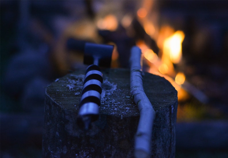 May include: A close-up of a black and white drill bit and a wooden stick resting on a tree stump. The background is blurred and shows a fire burning.
