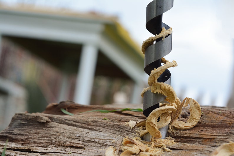 May include: A close-up of a drill bit with wood shavings wrapped around it. The drill bit is silver and black and is partially inserted into a piece of wood.