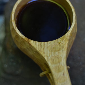 Op de afbeelding: Een houten beker met een donkerbruine vloeistof erin. De beker heeft een handvat en is gemaakt van lichtbruin hout.