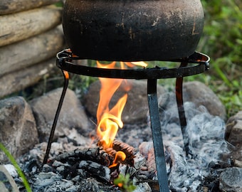 May include: A black clay pot with a lid sits on a metal tripod over a small fire. The fire is made of charcoal and wood and is burning brightly. The pot is surrounded by rocks and green grass.