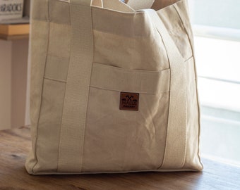 May include: A large, beige canvas tote bag with two handles and an exterior pocket. The bag has a rectangular shape and is sitting on a wooden table. A brown leather patch with a logo is visible on the front.