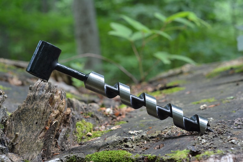 May include: A black and silver drill bit with a square head, lying on a piece of wood with moss.