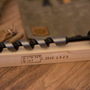 May include: A black and silver drill bit with a black handle and a ring on the end. The drill bit is resting on a wooden surface with the text "TR-347 HT 1 2020 1 3 7 5" engraved on the surface.