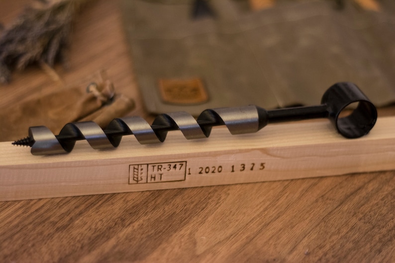 May include: A black and silver drill bit with a black handle and a ring on the end. The drill bit is resting on a wooden surface with the text "TR-347 HT 1 2020 1 3 7 5" engraved on the surface.