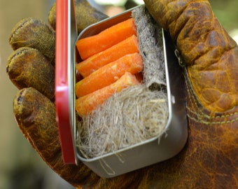 May include: A small metal tin with a red lid containing orange tinder sticks and a bundle of gray tinder. The tin is held in a brown leather glove.