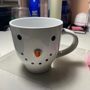 May include: White ceramic mug with a snowman face. The snowman has black eyes, a black mouth, a black hat, and an orange nose.
